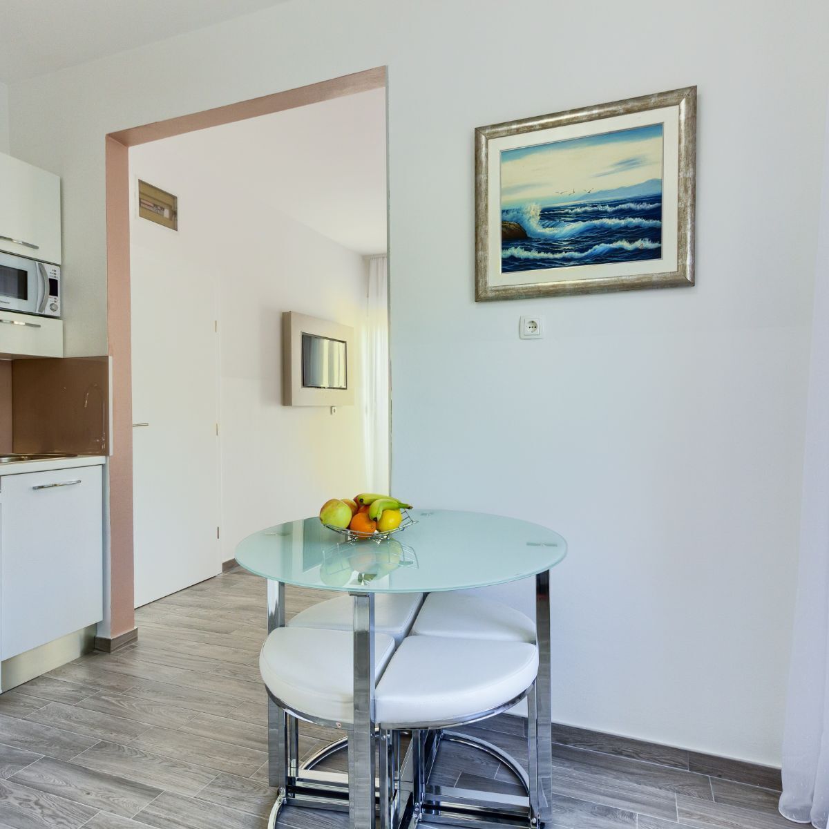 A modern kitchen with a microwave, a small refrigerator, and a glass table with a fruit bowl. There is a framed painting of a seascape on the wall.