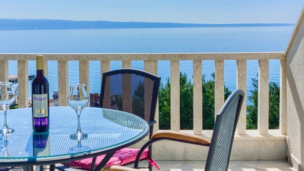 A balcony with a glass table, chairs, and a bottle of wine, overlooking a calm sea.