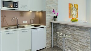 A modern kitchen with white cabinets, a built-in microwave, a stainless steel sink, and a dishwasher. There's a bar area with a stool and a potted orchid.