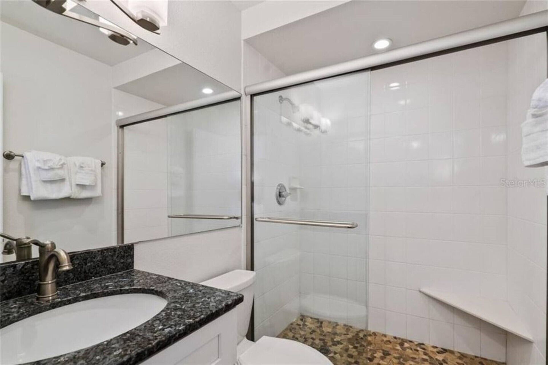 A modern bathroom with a granite countertop sink, a toilet, a glass shower enclosure, and a mirror.