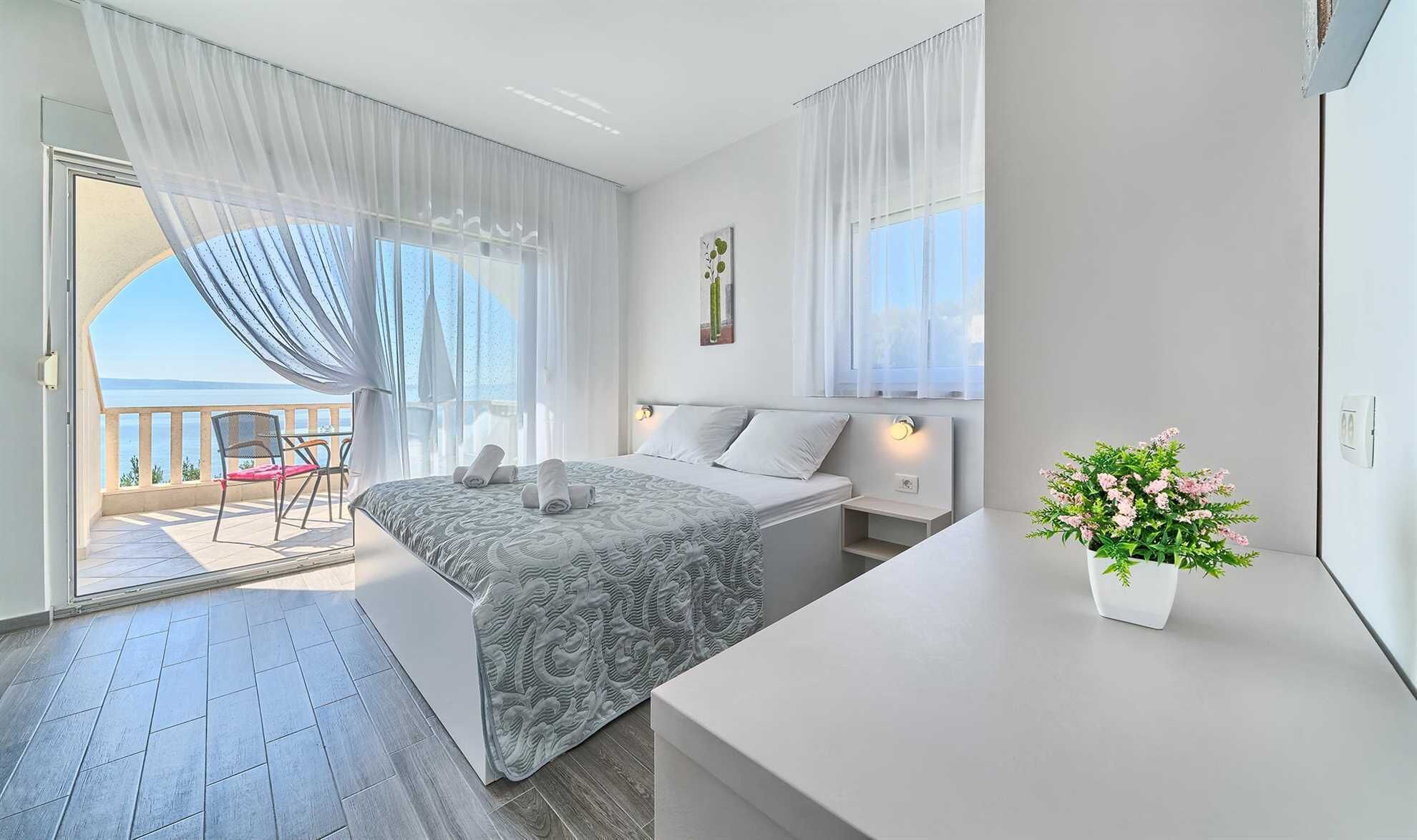 Un dormitorio con cama, mesita de noche, planta en maceta y balcón con silla.