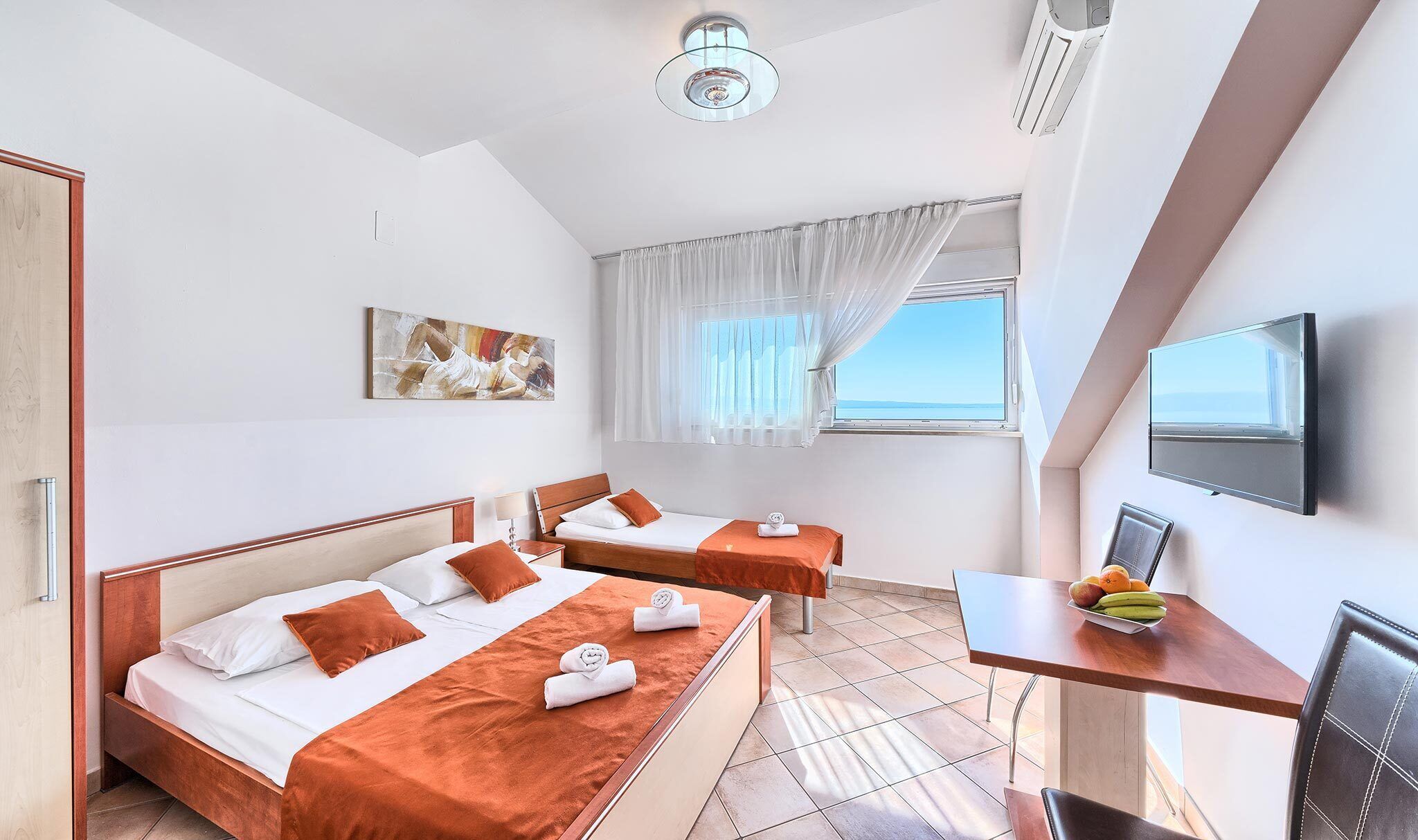 Quarto de hotel com duas camas, uma escrivaninha com televisão, um quadro e uma janela com vista.
