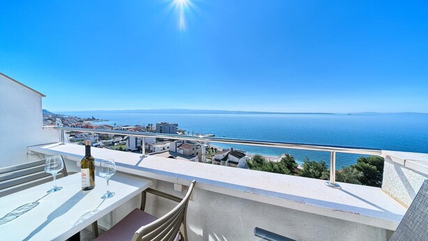 Een balkon met een tafel, twee wijnvaten en een fles wijn. In de verte een kustlandschap met gebouwen en de zee.