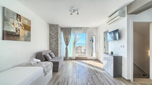 Een moderne woonkamer met een bank, een schilderij, een stenen muur, een schuifdeur naar een balkon en een keuken met een televisie.