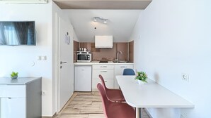 Een kleine keuken met een magnetron, spoelbak en kasten. Er staat een eettafel met stoelen en een klein plantje op de tafel.