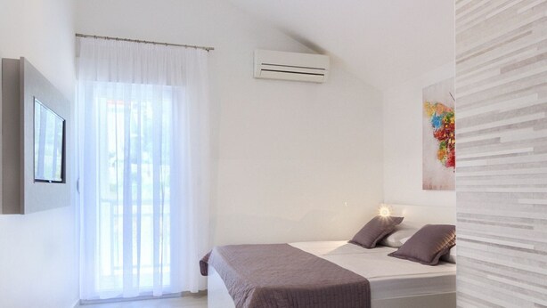 Un dormitorio moderno con una cama grande, aire acondicionado instalado, una ventana con cortinas translúcidas y un televisor montado en la pared.