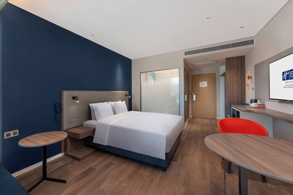 Une chambre d’hôtel moderne avec un grand lit, un bureau et une chaise. On y trouve une petite table et un téléphone sur la table de chevet.