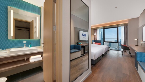 Habitación de hotel con baño, cama, escritorio y ventana grande con vistas a la ciudad.