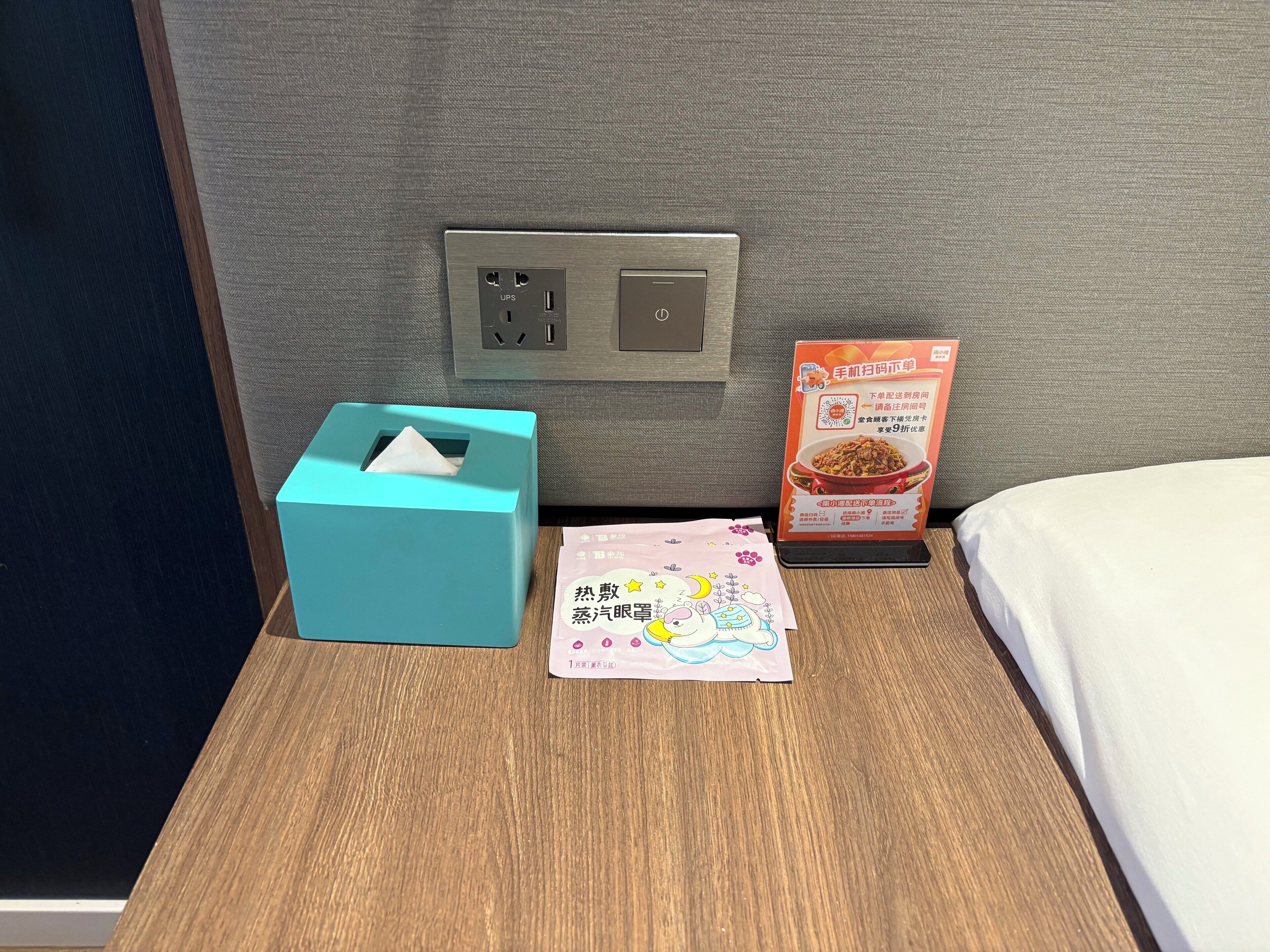Habitación de hotel con mesita de noche de madera, un dispensador de pañuelos color esmeralda, un paquete de aperitivos y un enchufe de pared.