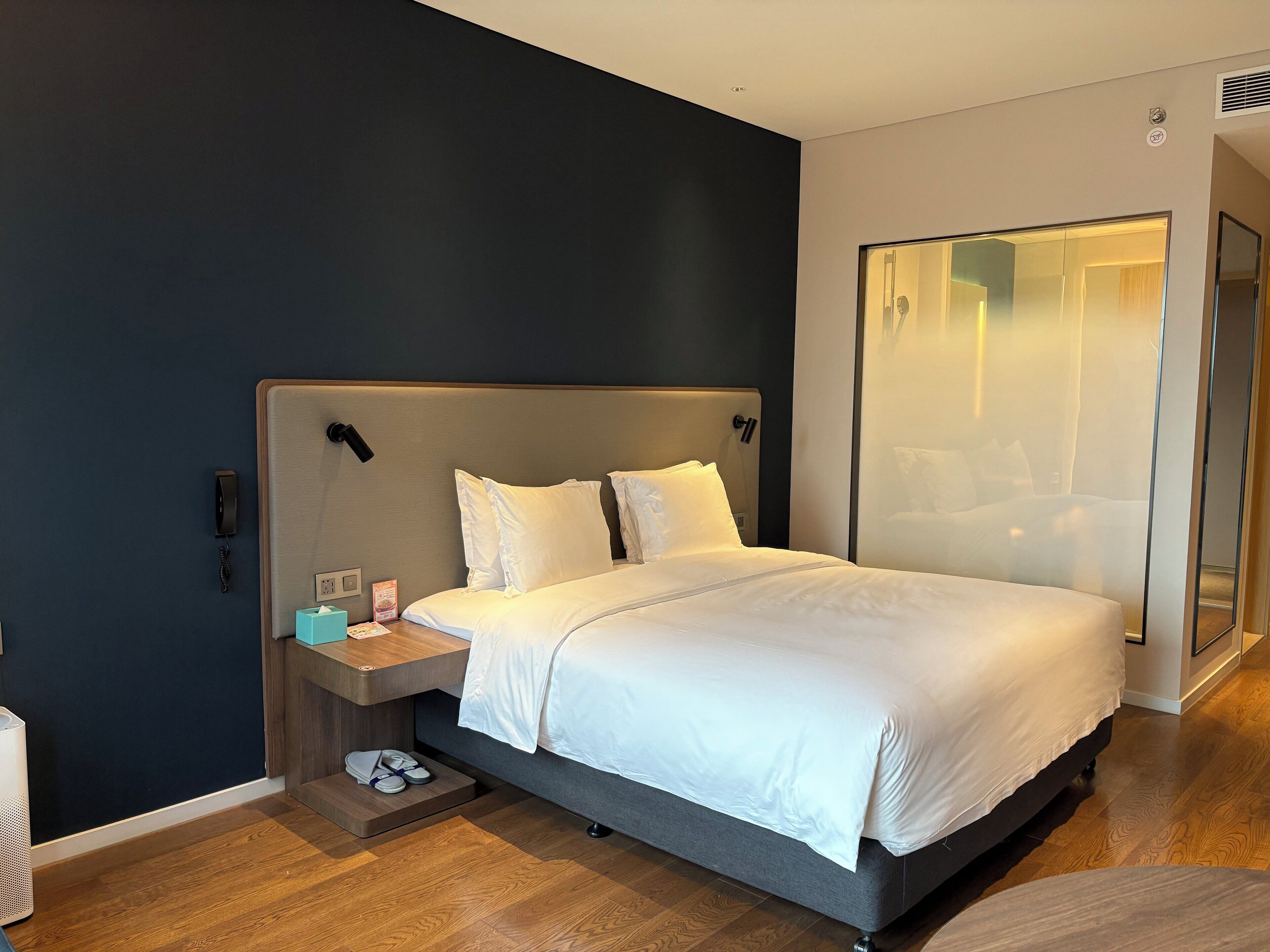 Una habitación de hotel moderna con una cama grande, mesitas de noche, una mesita auxiliar, una pared de vidrio y suelo de madera.