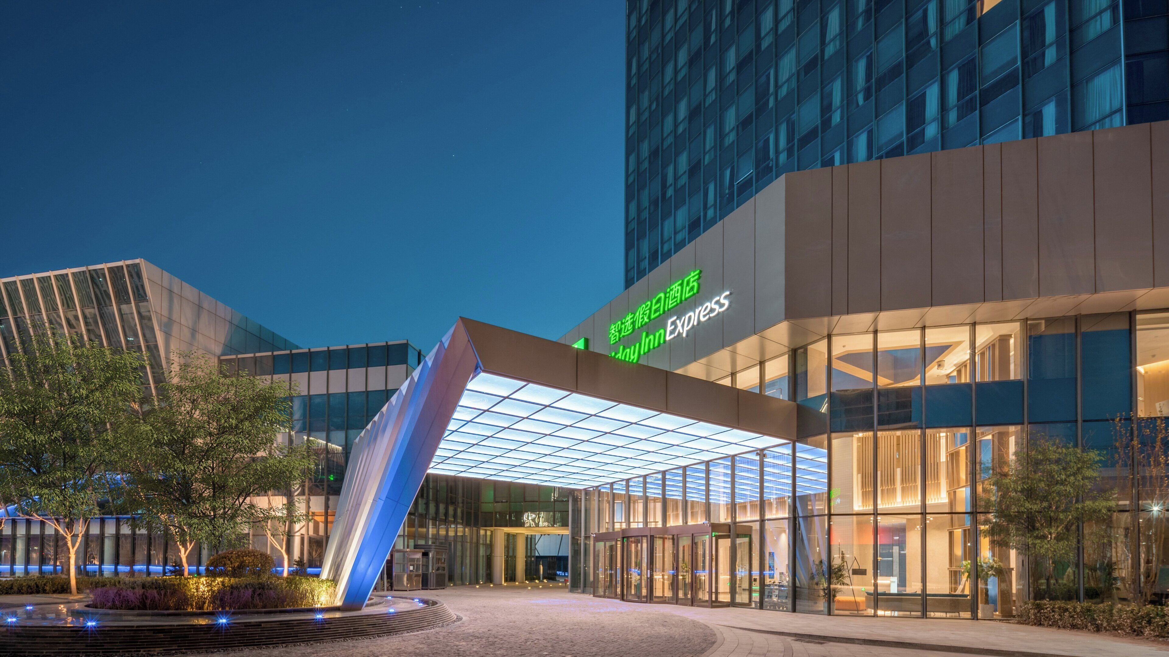 A modern hotel entrance with a glass canopy, a sign for 'Day Inn Express,' and a landscaped area with trees and a water feature.
