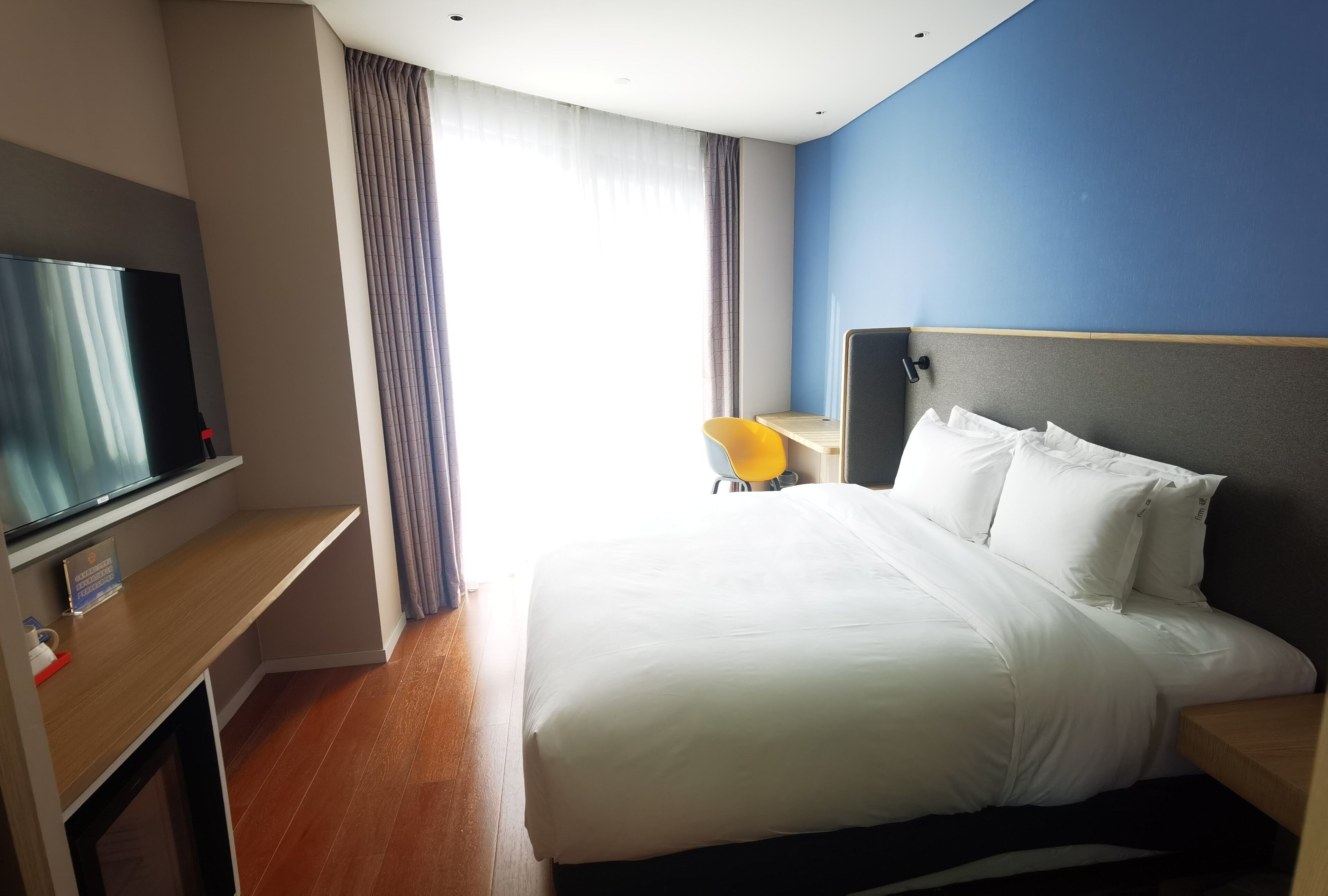 Habitación de hotel con cama, televisor sobre un mueble bajo, escritorio con silla y ventana con cortinas.