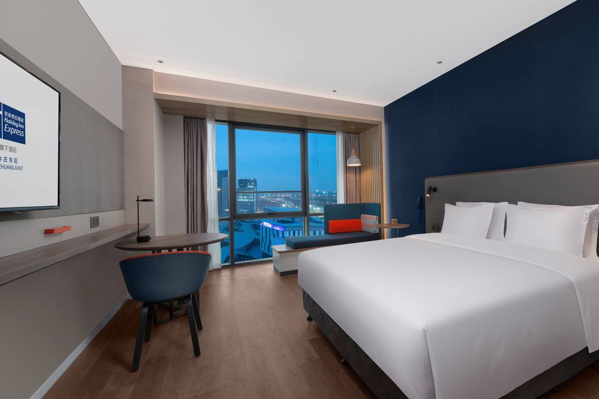 Une chambre d’hôtel avec un grand lit, un bureau, une chaise et une vue sur le paysage urbain grâce à de grandes fenêtres.