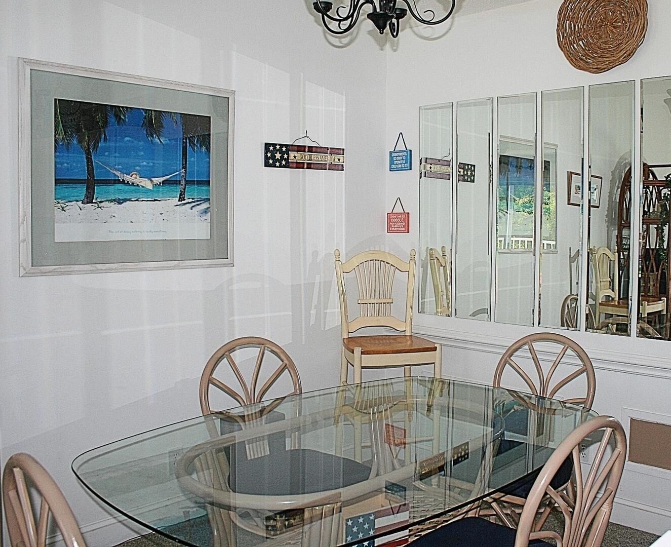 Un comedor con una mesa de vidrio, cuatro sillas de madera, una silla adicional y un cuadro enmarcado con una playa.