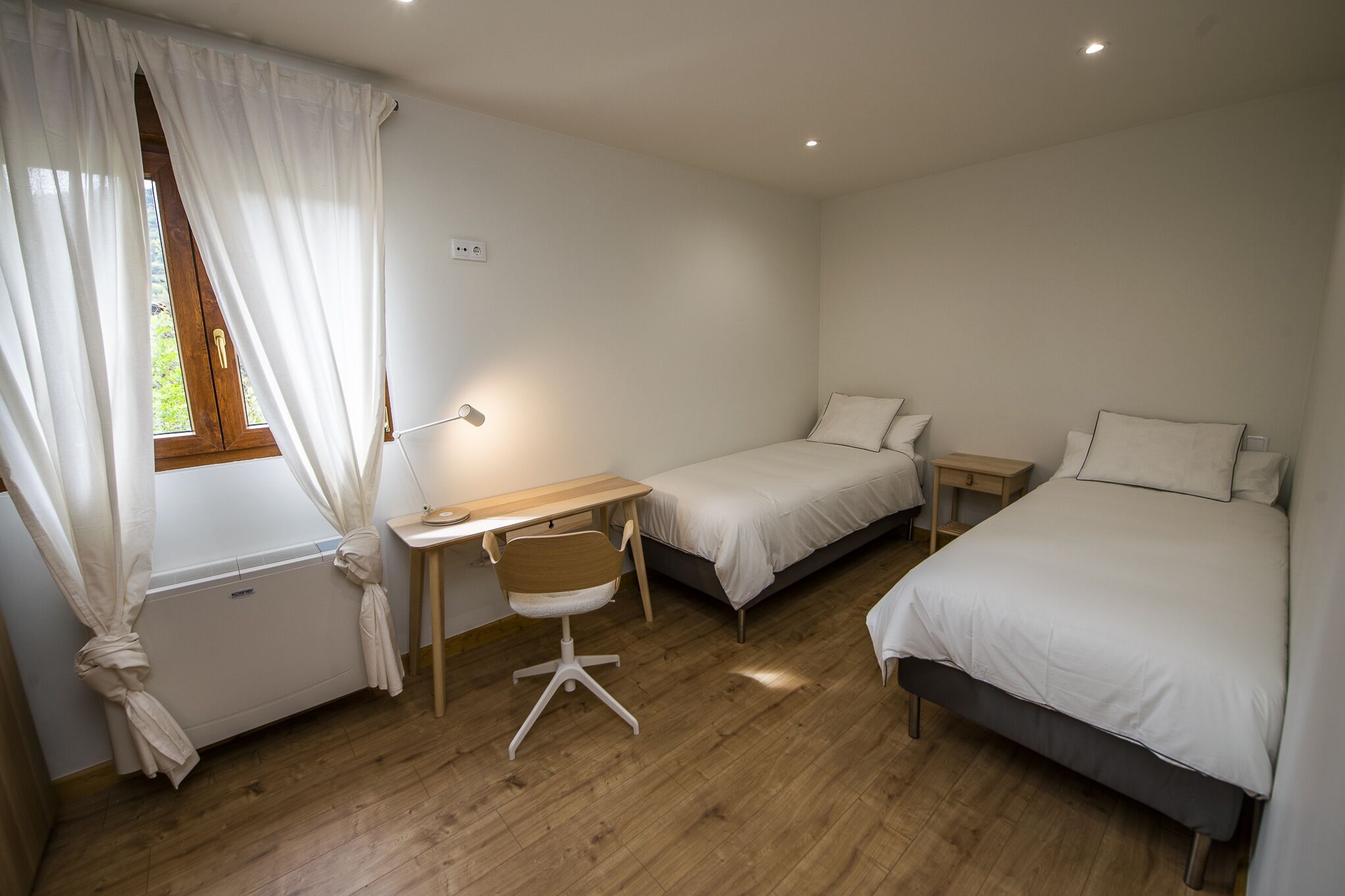 A bedroom with two single beds, a desk with a chair, a window with curtains, and a radiator.