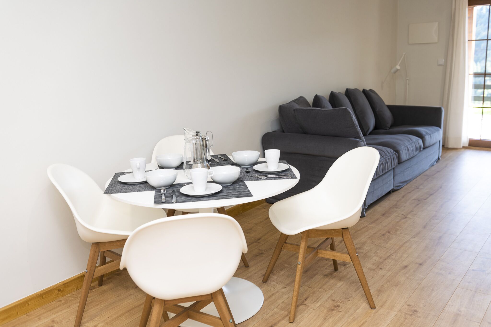 Ein Essbereich mit einem runden Tisch, gedeckt für vier Personen, weißen Stühlen und einem grauen Sofa im Hintergrund.