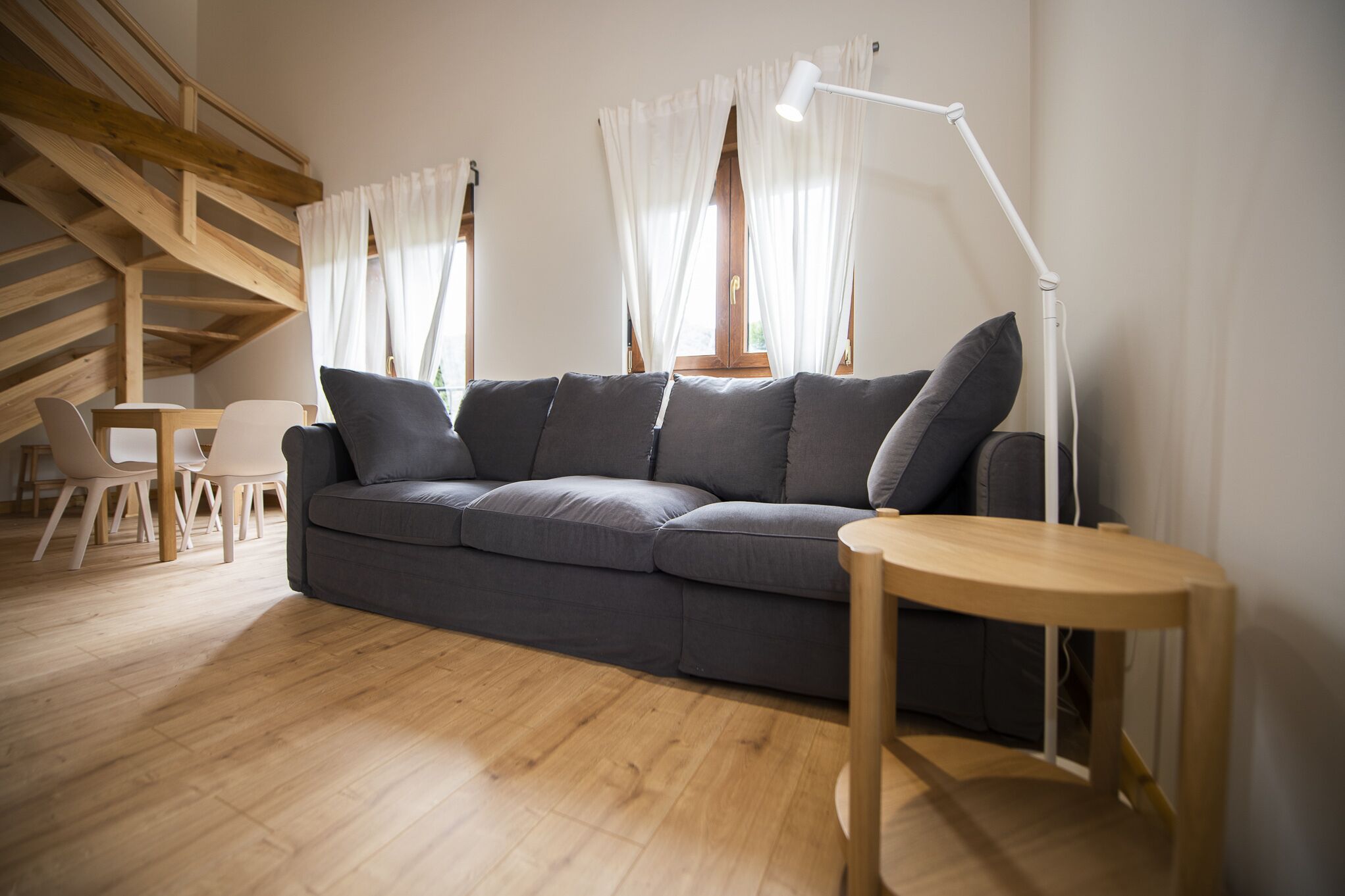 A modern living room with a grey sofa, wooden floor, white curtains, and a white floor lamp.