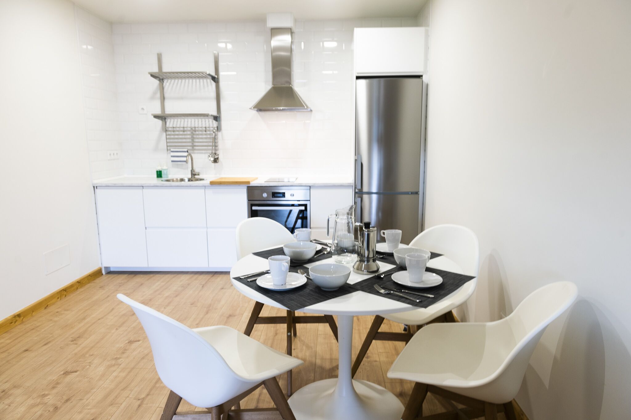 Una cocina moderna con armarios blancos, un refrigerador de acero inoxidable y un comedor con sillas blancas y mantel negro.