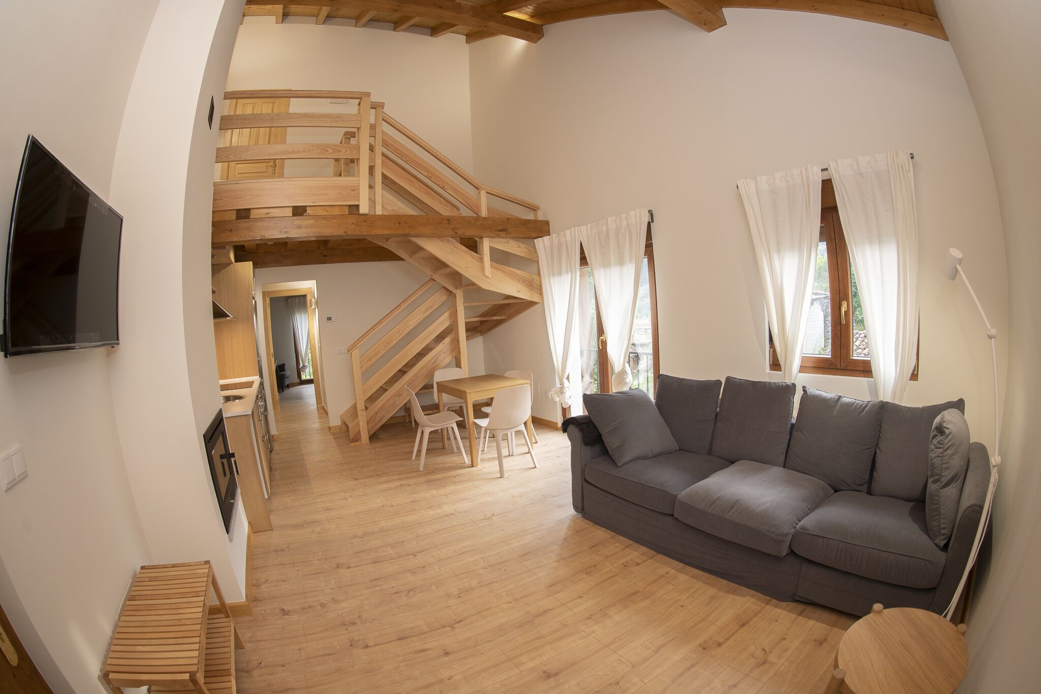 Uma sala ampla com escada de madeira, um sofá cinza e uma mesa de jantar com cadeiras.