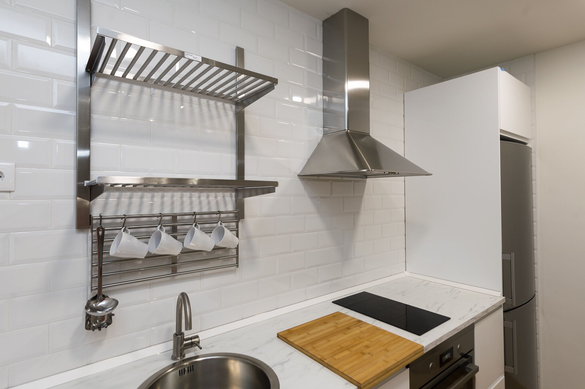 Una cocina moderna con azulejos blancos tipo metro, electrodomésticos de acero inoxidable, una tabla de cortar de madera y un escurreplatos metálico.