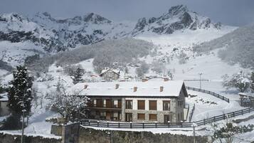 Un rifugio alpino imbiancato dalla neve, con mura in pietra e balconi in legno, circondato da alberi e montagne innevate.