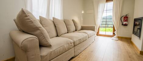 A beige sofa with multiple cushions, a wooden floor, a window with curtains, and a fire extinguisher on the wall.