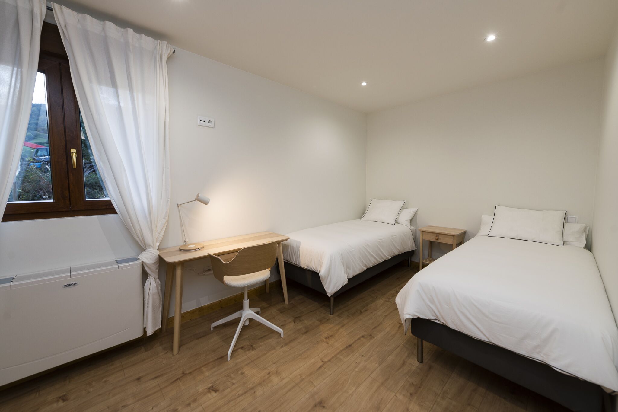 Un dormitorio con escritorio de madera, dos camas individuales, una mesita de noche y una ventana con cortinas.