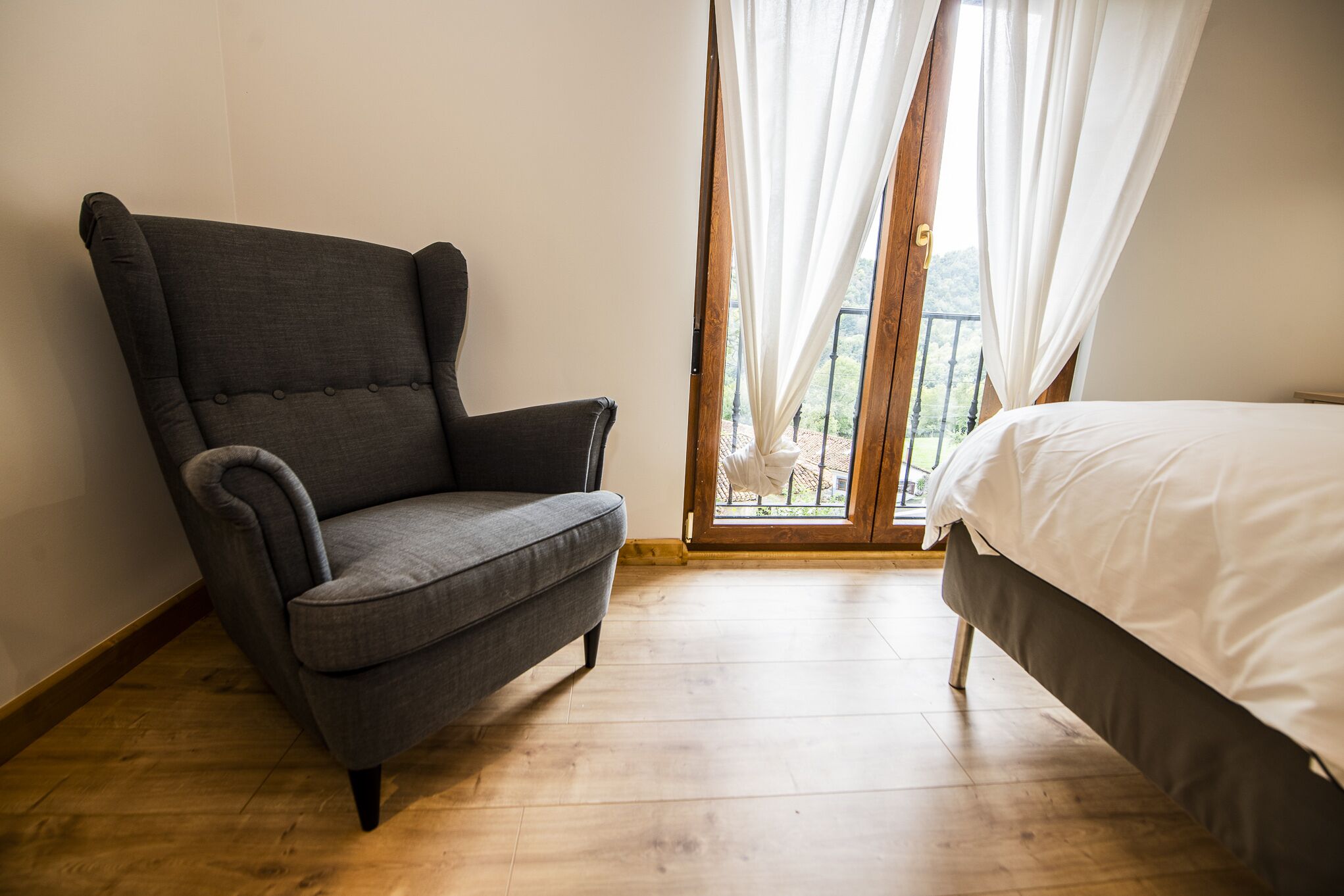A room with a grey armchair, a bed with white bedding, and a wooden floor.