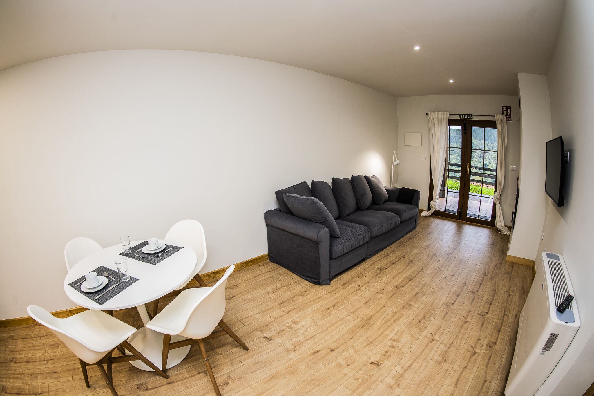 A modern living room with a dark grey sofa, white dining table with chairs, and a flat-screen TV mounted on the wall.