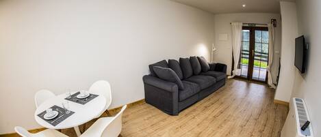 A modern living room with a dark grey sofa, white dining table with chairs, and a flat-screen TV mounted on the wall.