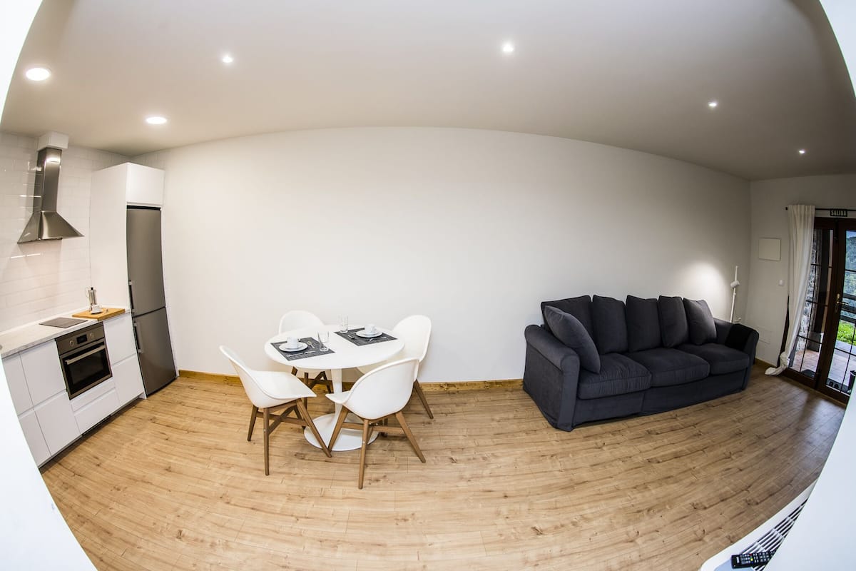 A modern kitchen with white cabinets, a dining table with white chairs, and a dark grey sofa in a living area with wooden flooring.