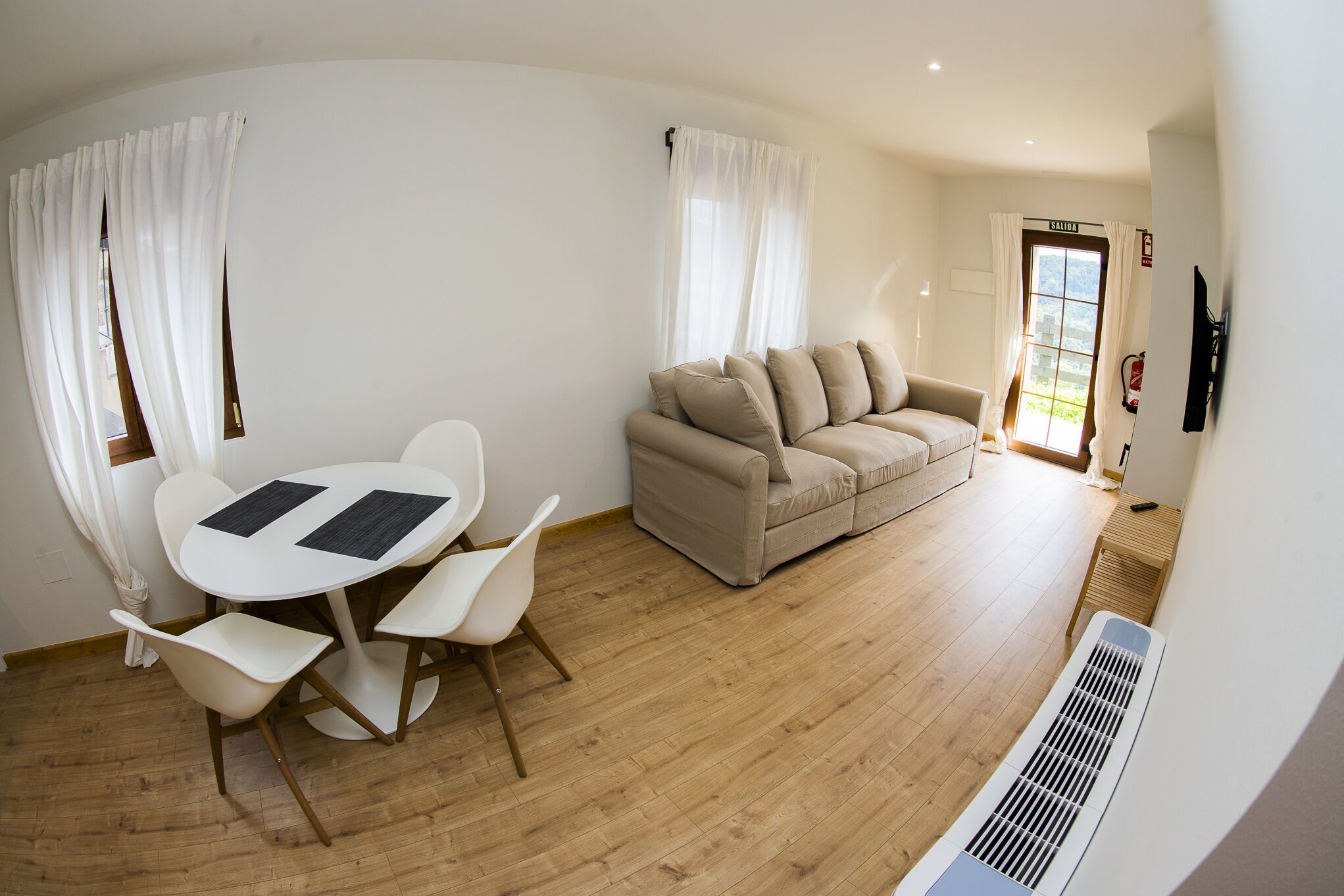 Una sala de estar moderna con un sofá color beige, una mesa de comedor blanca con cuatro sillas, y un ventanal grande con cortinas blancas.