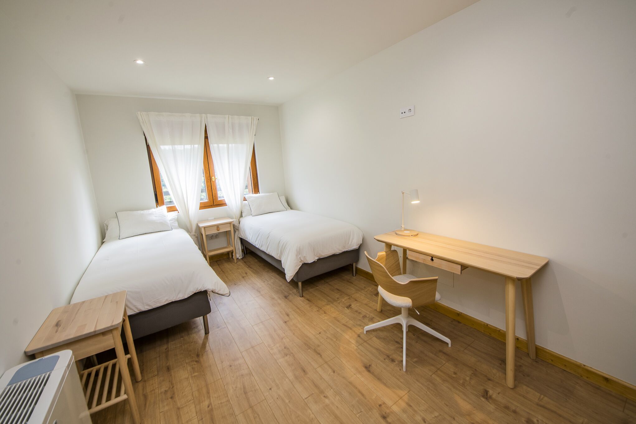 A small, well-lit room with two single beds, a wooden desk, and a chair.