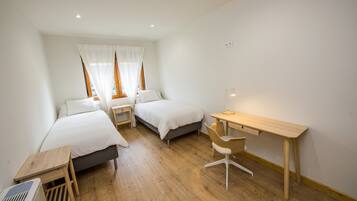 A small, well-lit room with two single beds, a wooden desk, and a chair.