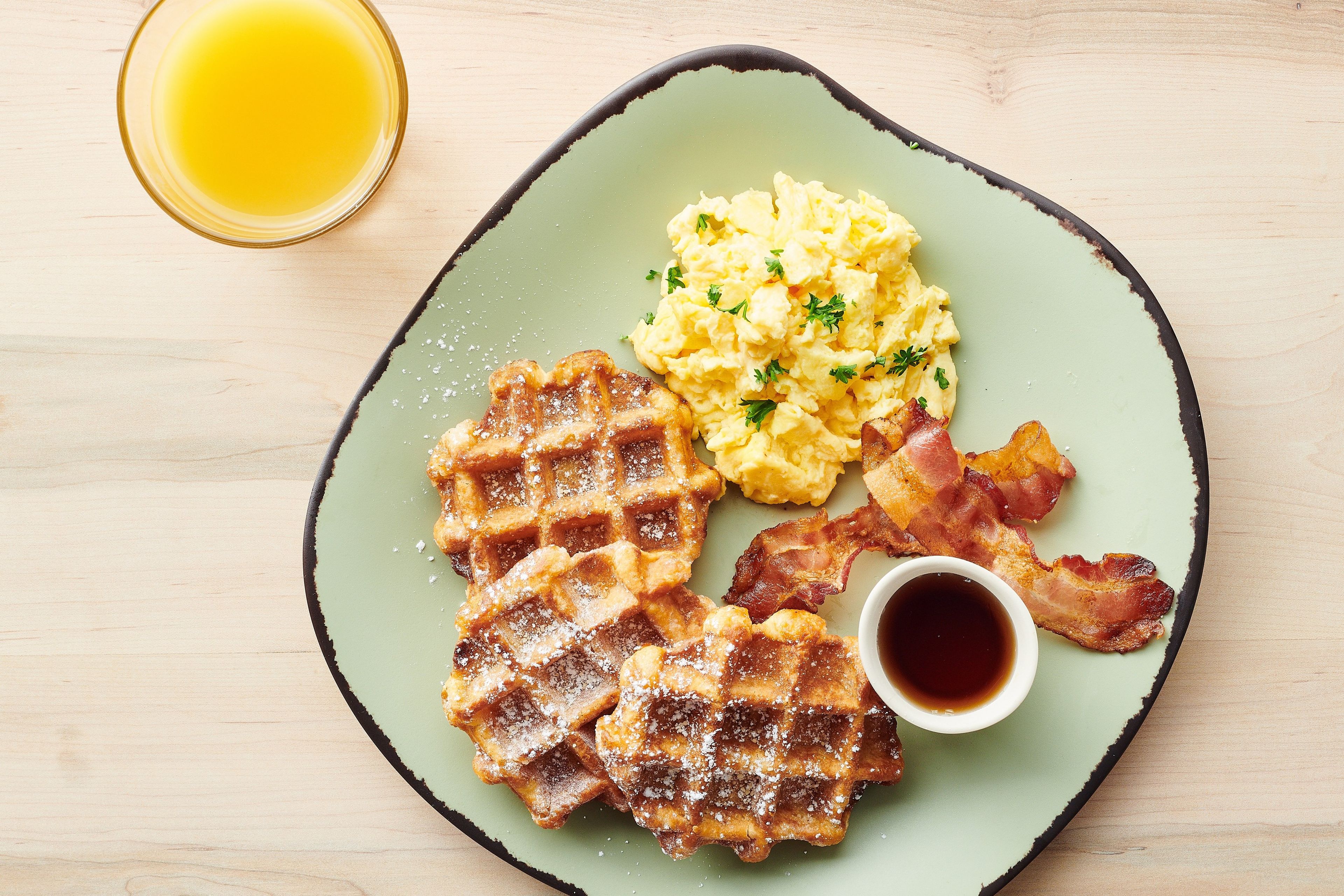A plate with waffles, scrambled eggs, bacon, and a cup of syrup.