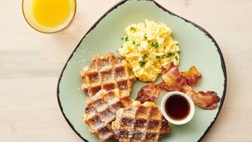 A plate with waffles, scrambled eggs, bacon, and a cup of syrup.