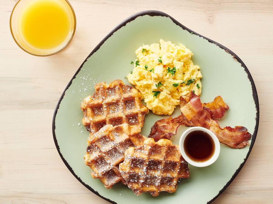 A plate with waffles, scrambled eggs, bacon, and a cup of syrup.