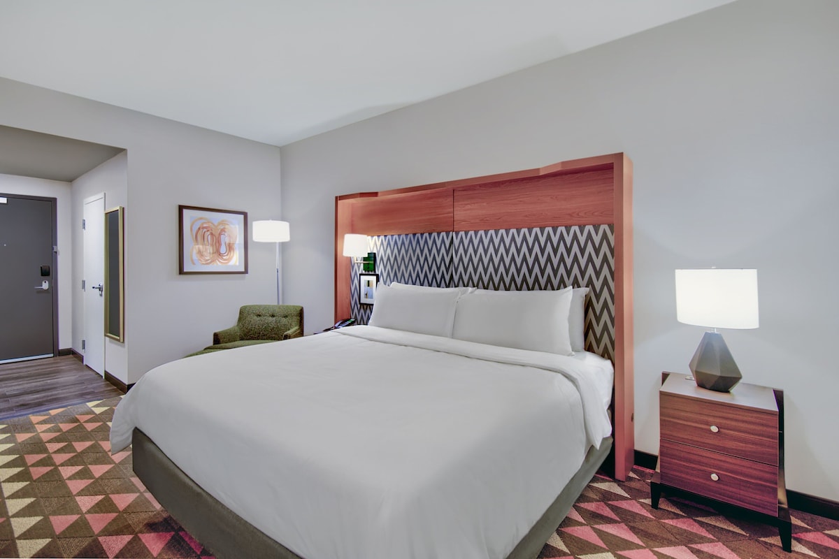 Une chambre d’hôtel avec un grand lit, une tête de lit en bois, une table de chevet avec une lampe, un fauteuil vert et une œuvre d’art accrochée au mur.