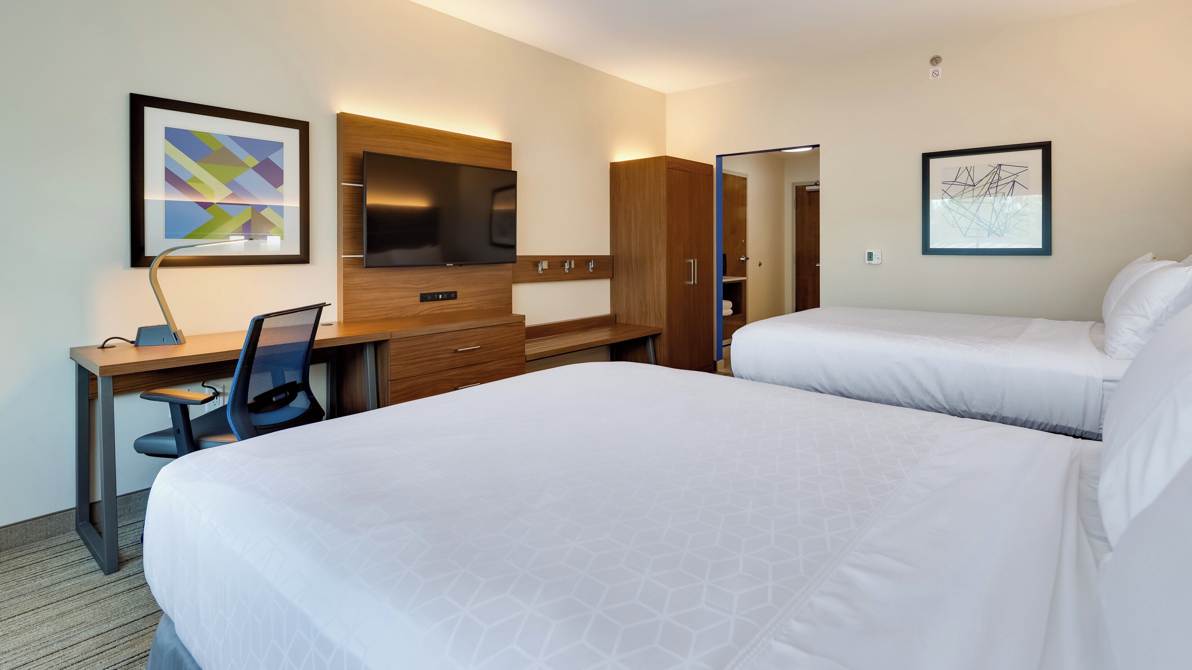 Habitación de hotel con cama, escritorio, silla, televisor, armario y obras de arte en las paredes.
