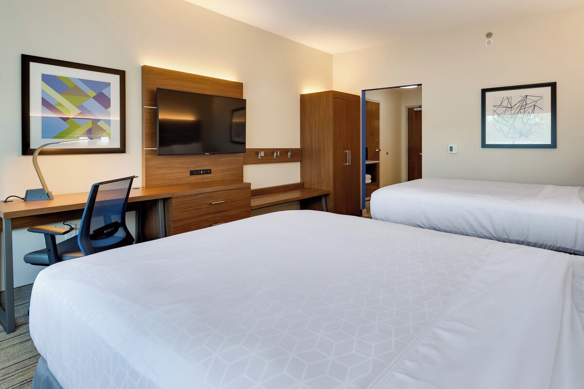 A hotel room with two beds, a desk with a chair, a TV, and artwork on the walls.