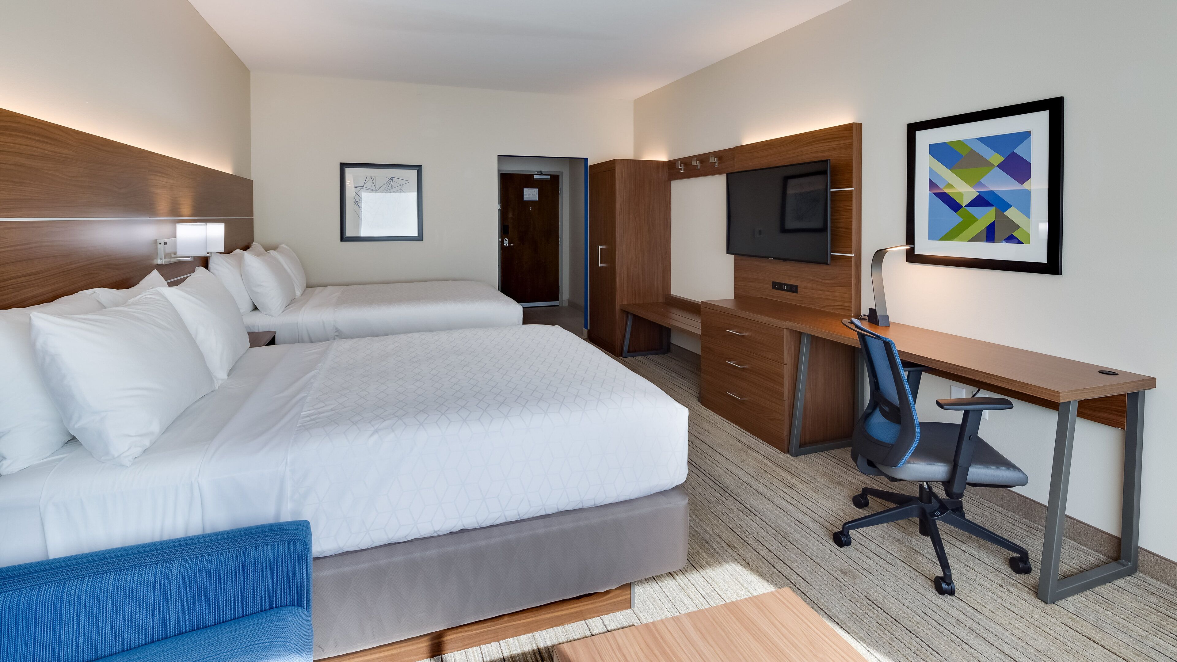 A hotel room with two beds, a desk, a chair, a TV, and artwork on the wall.