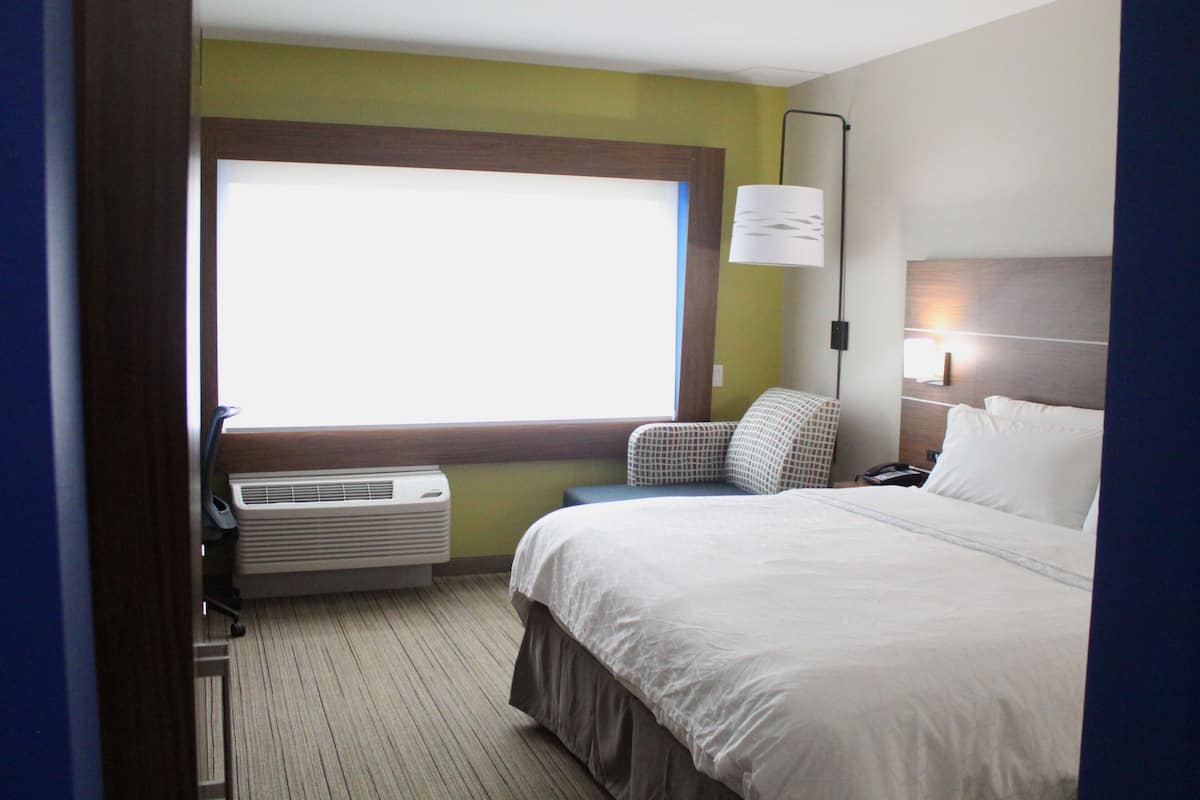 Une chambre d’hôtel avec une grande fenêtre, un lit, une chaise, un bureau et une lampe.