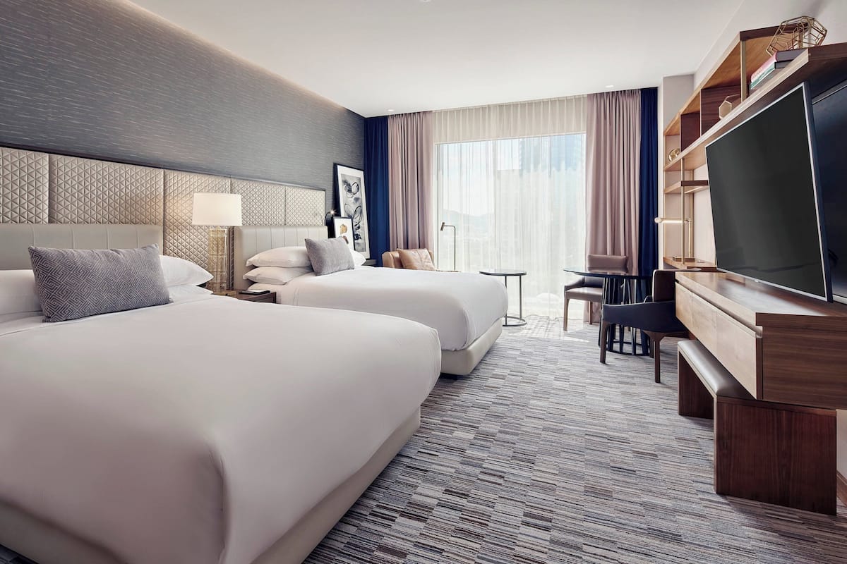 Une chambre d’hôtel avec deux lits, un bureau, une télévision et une grande fenêtre avec des rideaux.