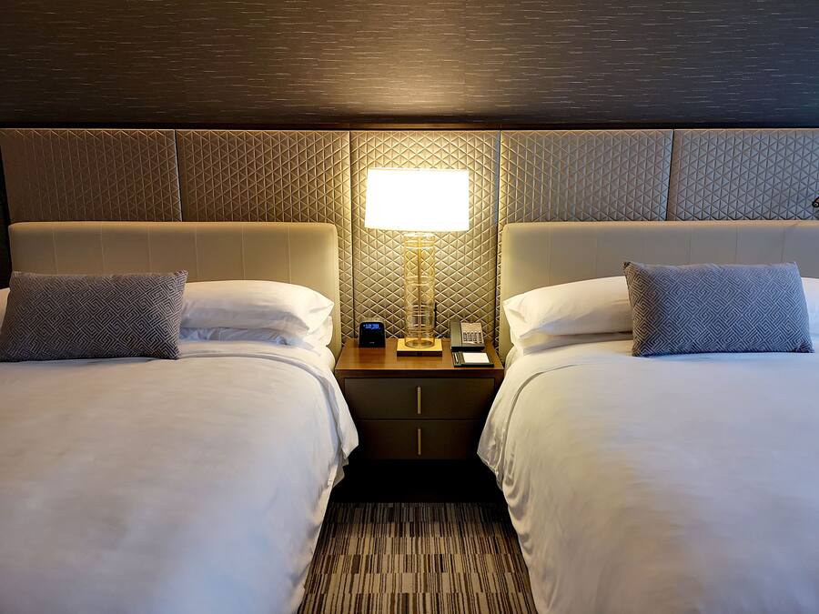 Habitación de hotel con dos camas, una mesita de noche con lámpara, reloj y teléfono.