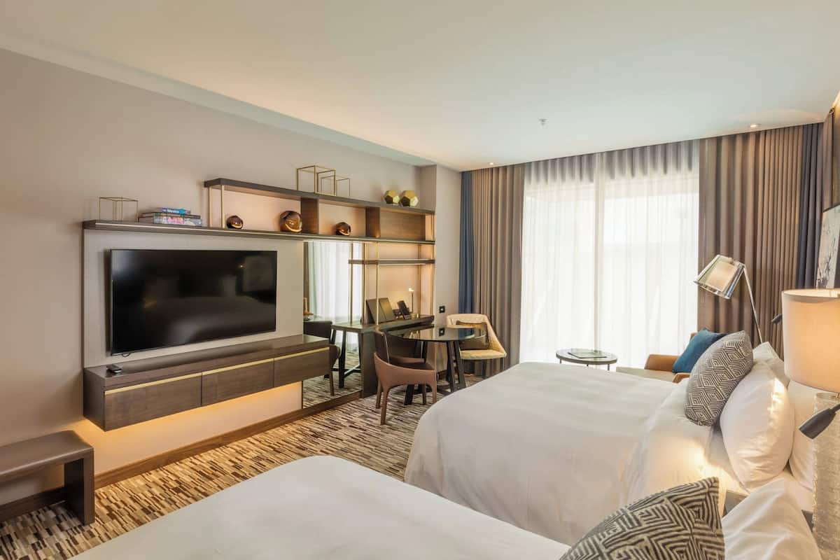 Une chambre d’hôtel dotée d’une grande télévision à écran plat, d’un bureau avec une chaise, d’un lit avec du linge de lit blanc et d’une grande fenêtre avec des rideaux.