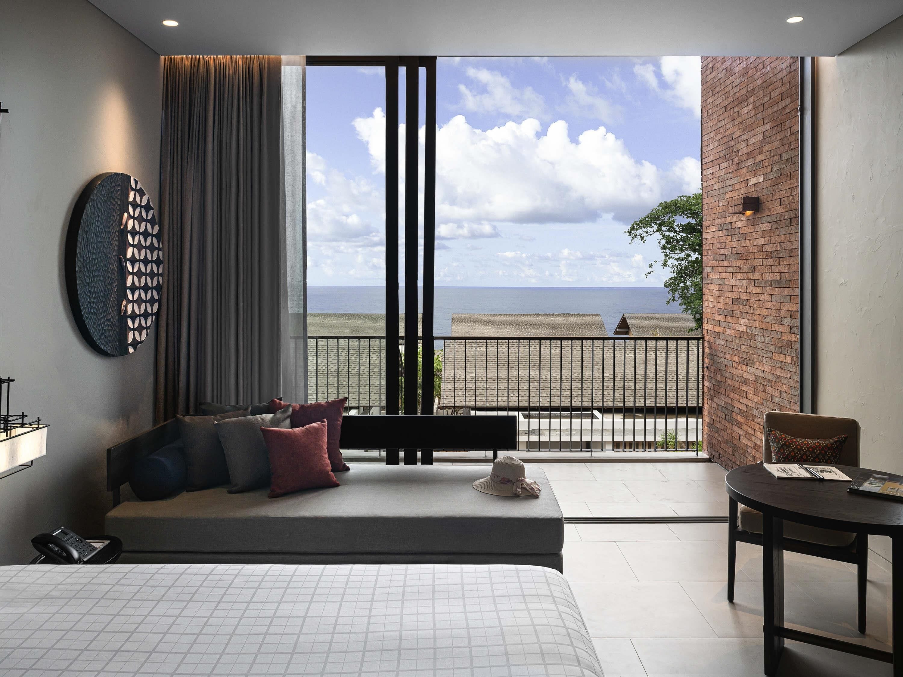 A modern hotel room with a large bed, a seating area with a view of the beach, and a balcony with a brick wall.