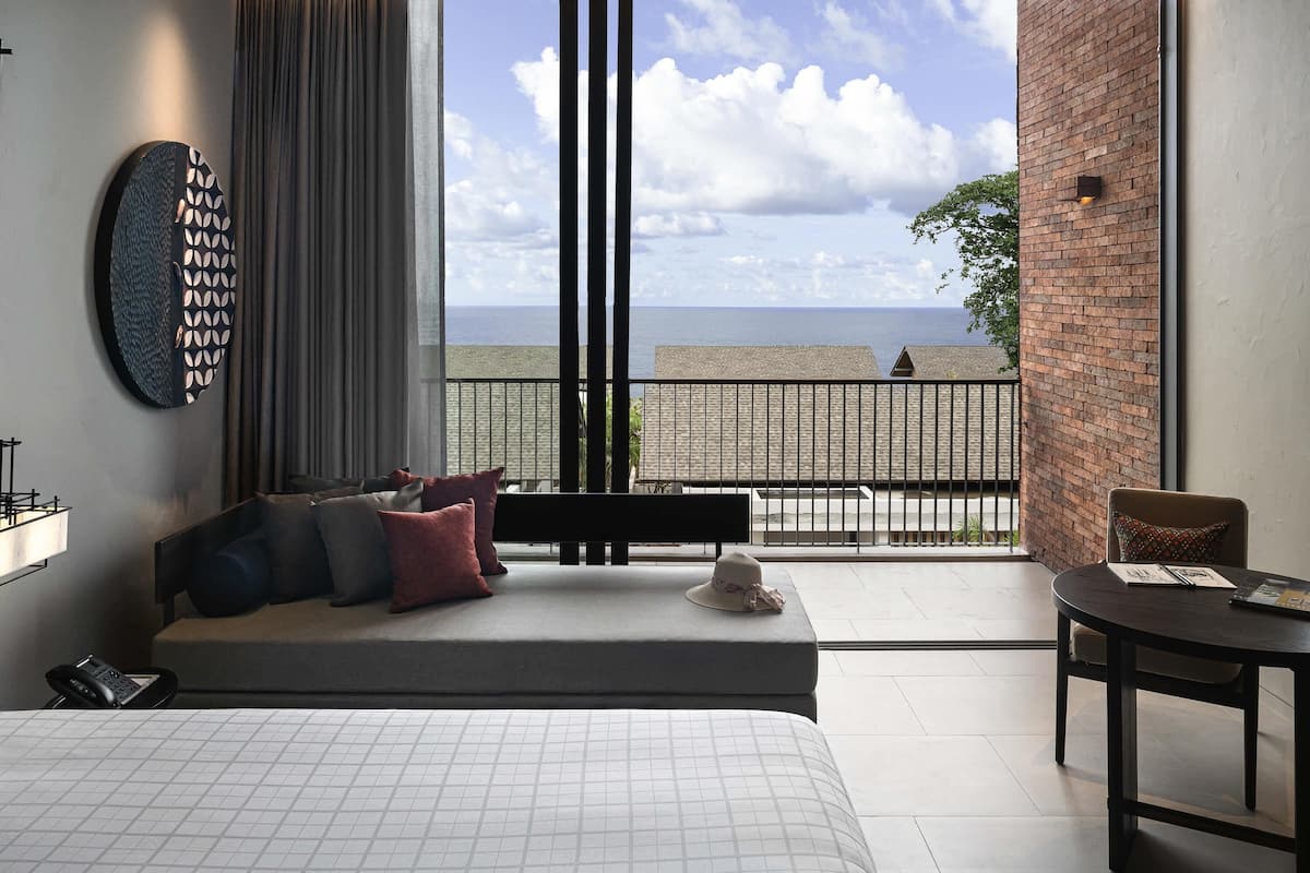 A modern hotel room with a large bed, a seating area with a view of the beach, and a balcony with a brick wall.