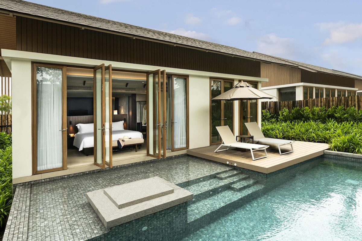 A modern hotel room with a glass door leading to a pool area, including a bed, a bathroom, and a seating area with a poolside umbrella.