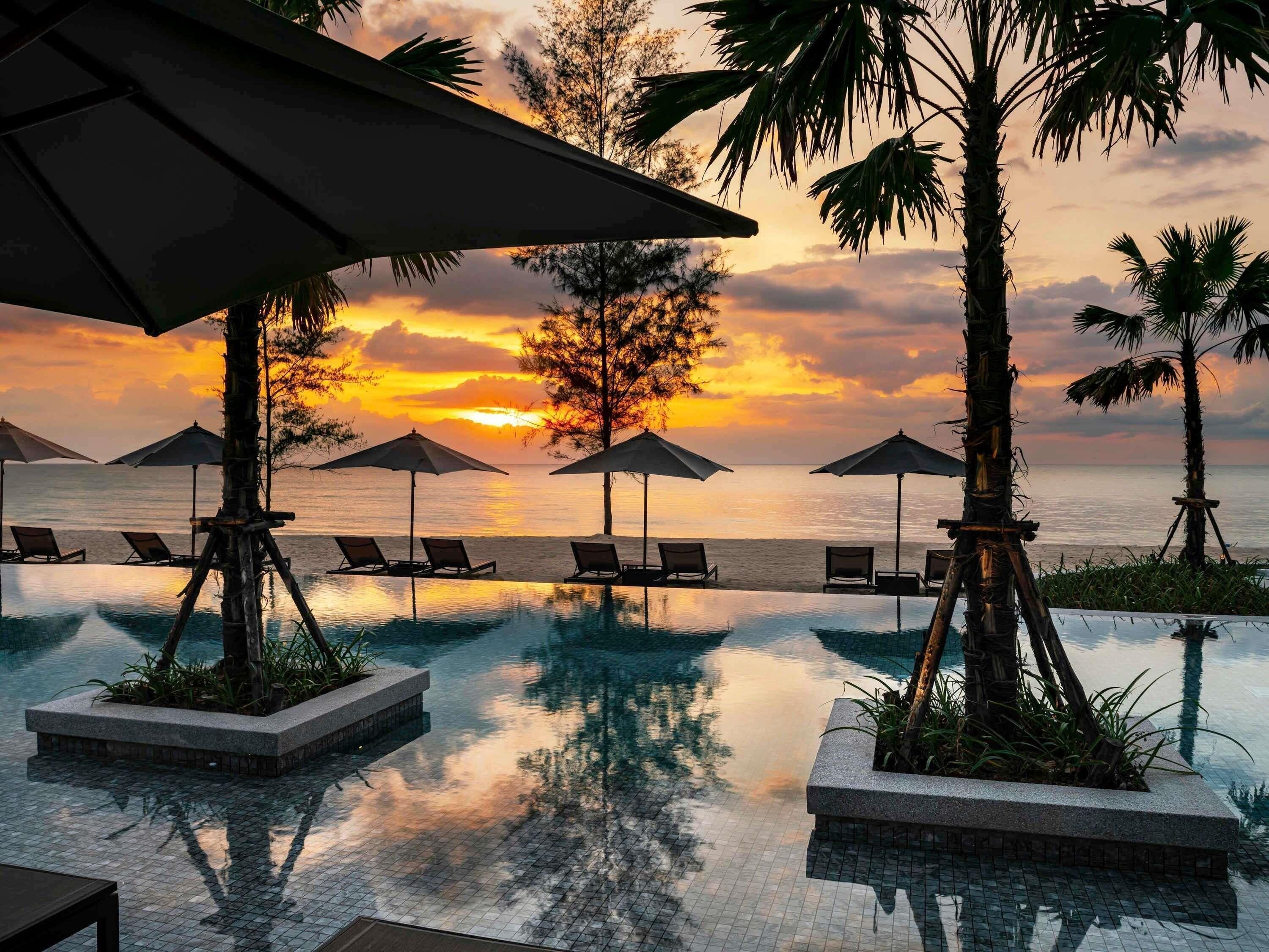 An infinity pool with lounge chairs and umbrellas, overlooking a sunset over the ocean.