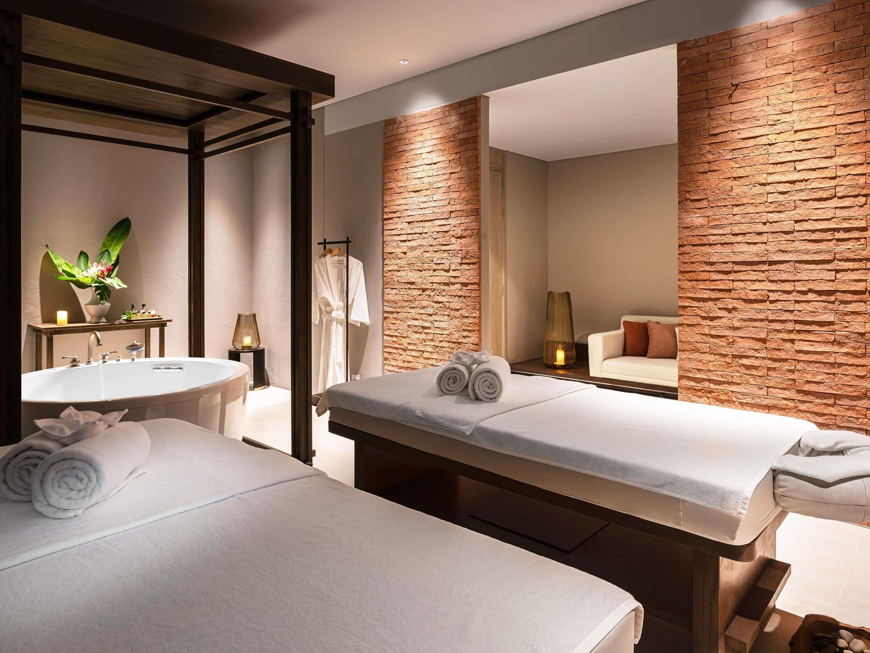 A modern hotel room with a large mirror, a massage table, a bed, a round sink, and a small plant.